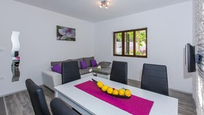 A modern living room with a white sofa, black chairs, a dining table with a pink placemat, and a window with a view of greenery. - Villa Roza- Two-Bedroom Apartment (Mokosica)