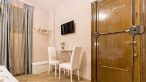 Une chambre d’hôtel avec une porte en bois portant l’inscription « Petites suites », un lit, une petite table avec des chaises, un téléviseur fixé au mur et une fenêtre avec des rideaux légers.