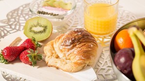 Une table blanche, agrémentée d’une nappe décorative, est dressée pour le petit-déjeuner : on y trouve un croissant, des fraises, un kiwi, un saladier de fruits et un verre de jus d’orange.