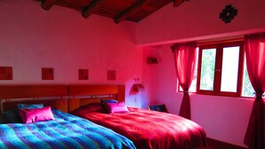 Ein Zimmer mit zwei Betten, einer Holzbalkendecke, einem Fenster mit roten Vorhängen und einer Wand mit gerahmten Bildern.