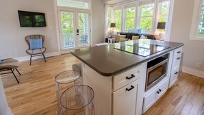 A modern kitchen with a large island, white cabinetry, and a built-in oven. There are two bar stools and a wicker chair. - Luxury Beachwalk Resort House Closest to Beach includes resort amenities (Michigan City)