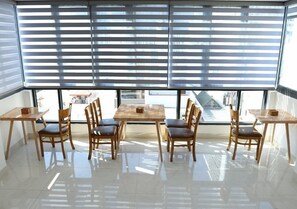 A dining area with a large table and chairs, large windows with blinds, and a view of the cityscape.
