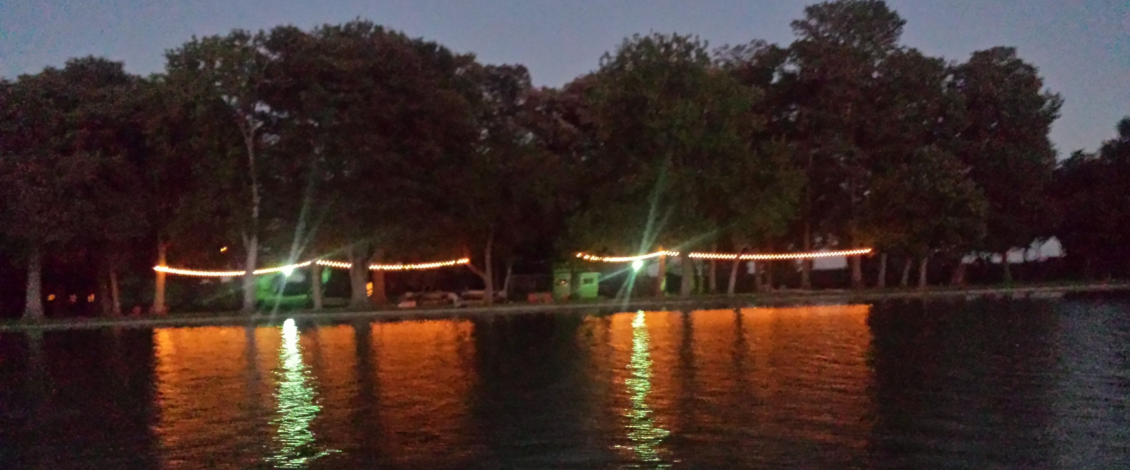 Vista nocturna de un parque con árboles y un cuerpo de agua que refleja luces.