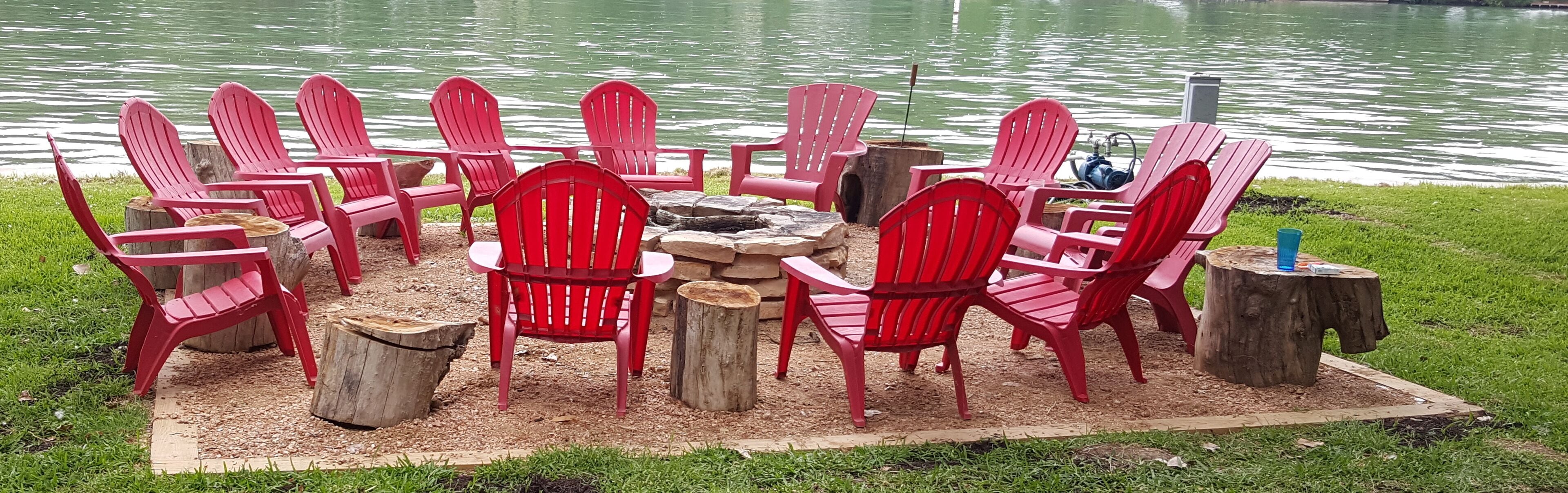 Un espace de détente circulaire, avec des chaises rouges disposées autour d’un foyer central, aménagé sur une pelouse près d’une étendue d’eau.
