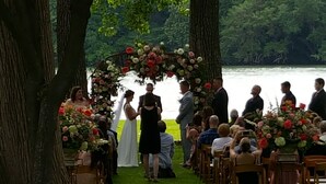 A wedding ceremony outdoors with a floral arch and guests seated on wooden chairs.