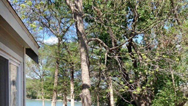 A deck with a glass table and chairs, overlooking a lake and trees.