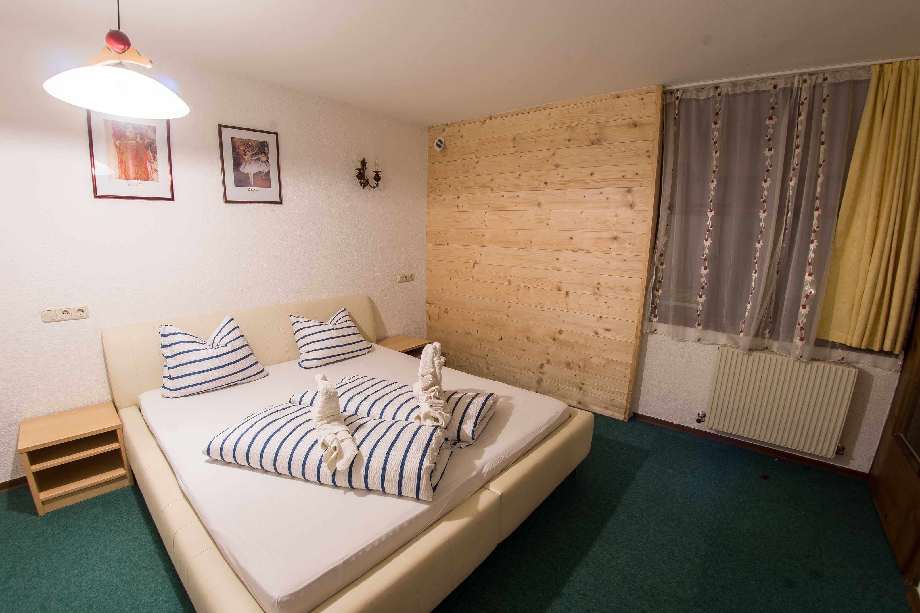 Een slaapkamer met een bed, kussens, een nachtkastje, een houten hoofdbord, een wandlamp, ingelijste foto's en een raam met gordijnen.