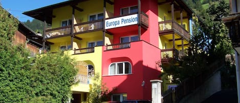 A multi-story building with balconies, labeled 'Europa Pension,' featuring a red and yellow facade, parked cars, and a mountainous backdrop.