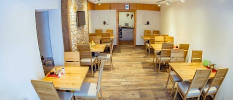 A dining area with wooden tables and chairs, a stone wall, and a kitchen visible through an open door.