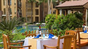 Comedor con sillas y mesas de madera, piscina al fondo y un edificio con balcones.