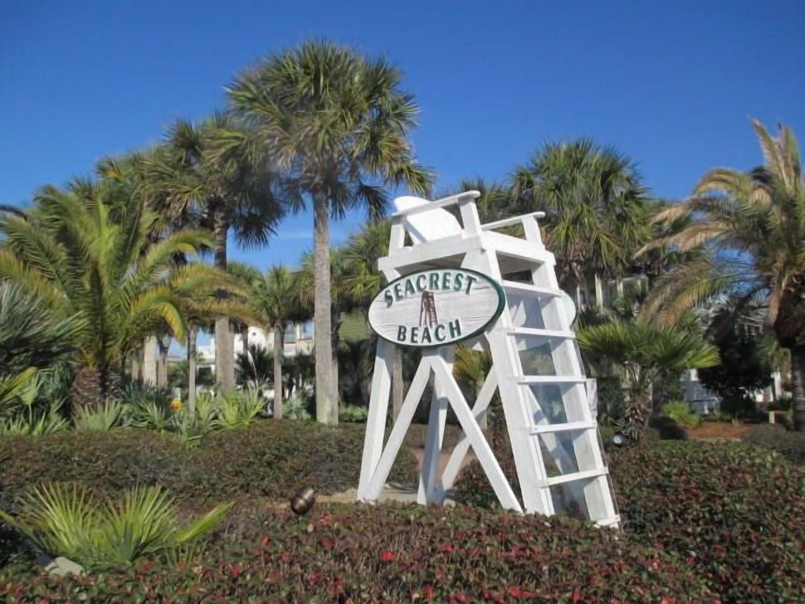 Schild für Seacrest Beach mit einem weißen, geometrischen Design und Palmen im Hintergrund.
