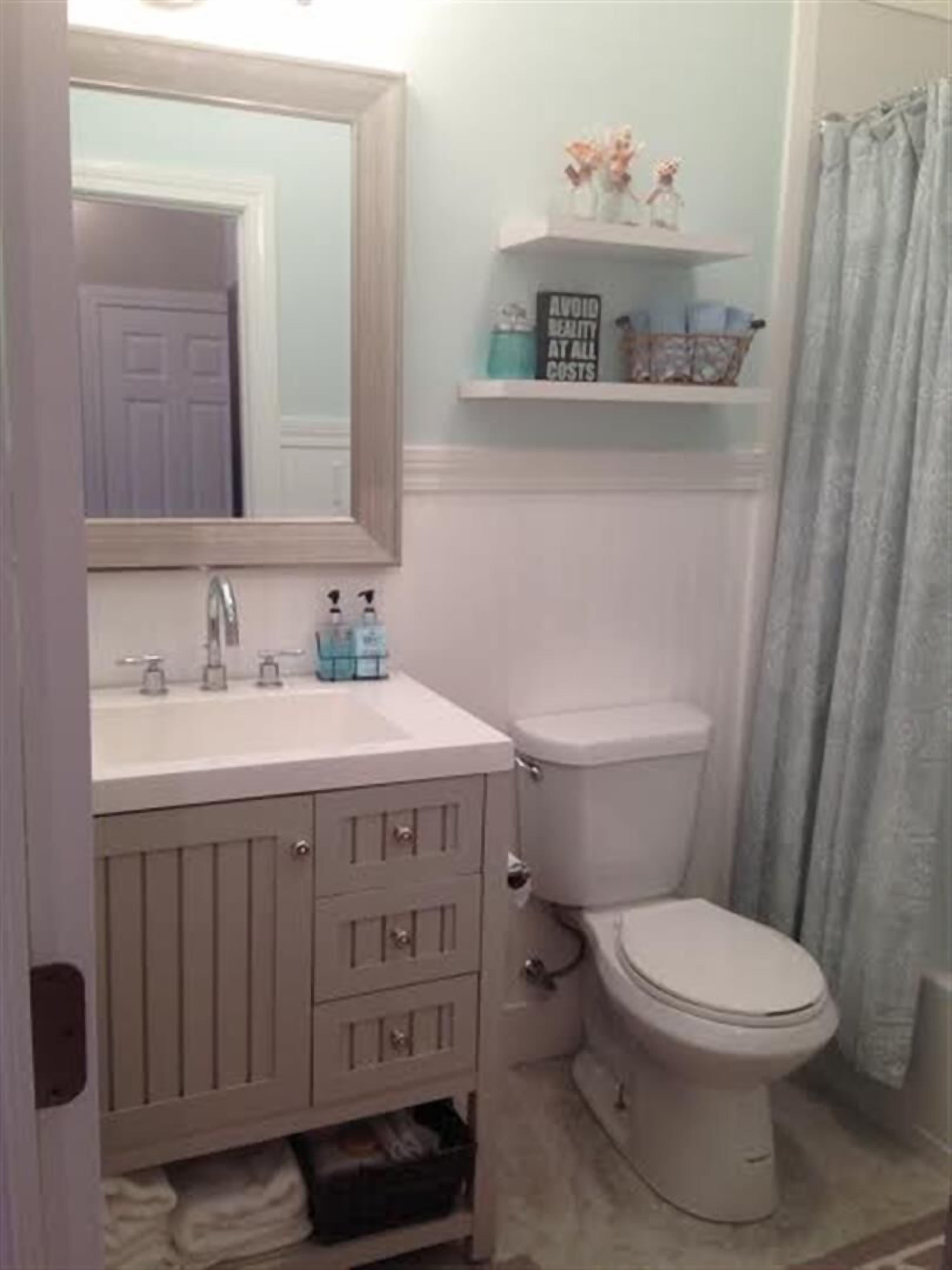 Un baño con lavamanos, espejo y inodoro blancos. Hay un estante sobre el inodoro con objetos decorativos.