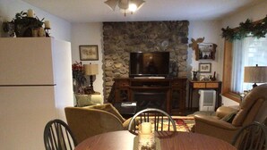 Una sala de estar acogedora con pared de piedra, televisor, chimenea y comedor con mesa redonda y sillas.