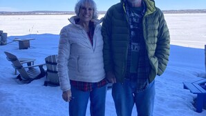 Dos personas de pie en un espacio exterior nevado con cielo despejado. Se ven sillas Adirondack y una mesa al fondo.