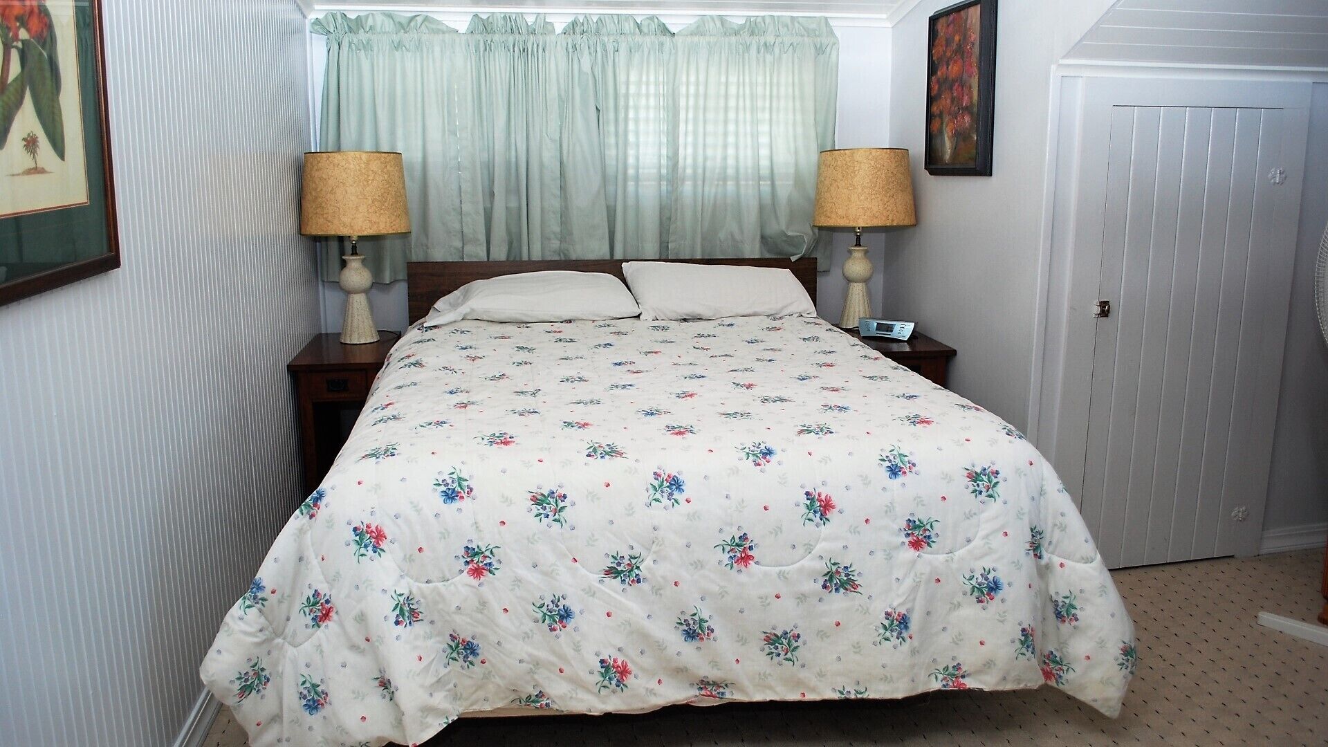 Un dormitorio con una colcha estampada de flores, dos lámparas de noche, una mesita de noche, un cuadro enmarcado en la pared y una puerta.