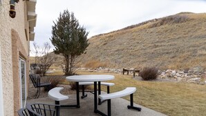 An outdoor patio with a round table and benches, a building with a window, and a hillside in the background. - Paradise Canyon Golf Resort - Luxury Condo M409 (Lethbridge)