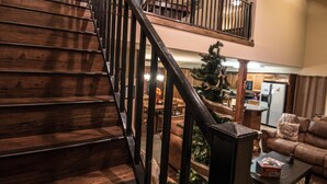 Una escalera de madera con barandilla negra que conduce a un balcón en el segundo piso. Se observa un árbol de Navidad al fondo, y en primer plano un área de estar acogedora con un sofá y una mesa de centro.
