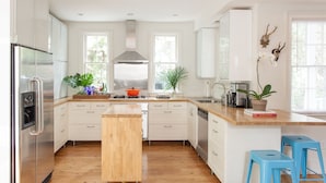 A modern kitchen with white cabinets, a stainless steel refrigerator, a wooden island, and blue bar stools. - Palm House- Prime Setting and 5 Stars (Charleston)