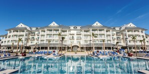A large hotel with a swimming pool, multiple balconies, and palm trees.