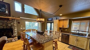 A spacious kitchen with a large island, wooden chairs, a stone fireplace, and a view of the outdoors.