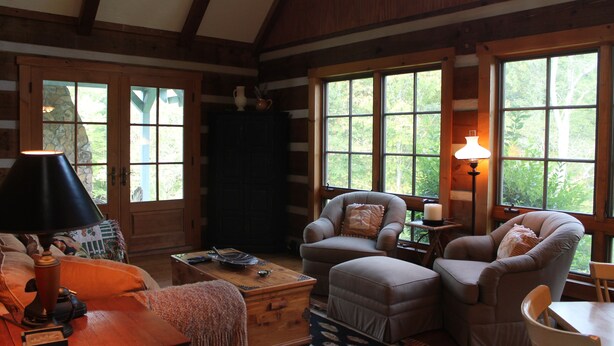 A cozy cabin interior with a fireplace, wooden walls, large windows, and comfortable seating. - Blue Ridge Mountain 'Solitude' on The Little River (Sparta)