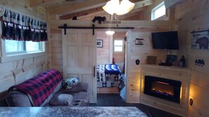 A cozy cabin interior with a sofa, a fireplace, a television, and a door leading to another room. - Cedar Bear Cabin - Cozy Mountain View Couples Retreat (Franklin)