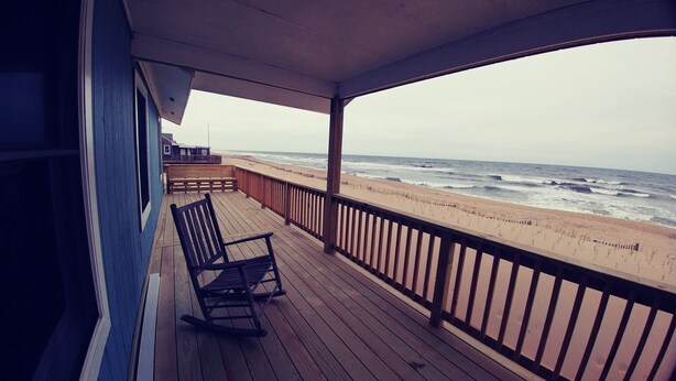 Une terrasse en bois avec un fauteuil à bascule et un banc, donnant sur une plage où l’on peut admirer les vagues.