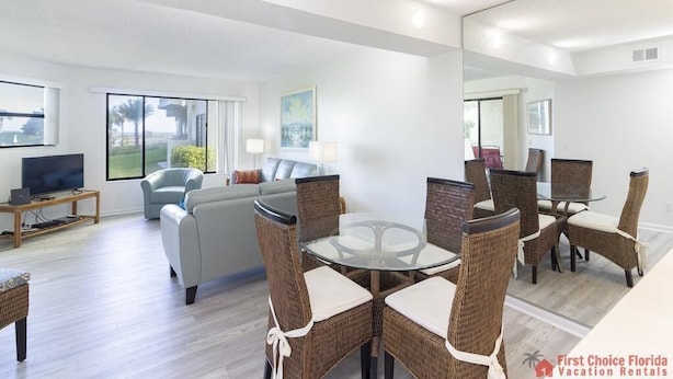 A modern living room with a glass-top dining table surrounded by wicker chairs, a grey sofa, and a flat-screen TV on a wooden stand.