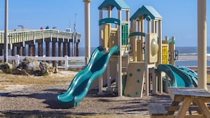 A playground with slides and climbing structures, a wooden bench, and a white fence in the background.
