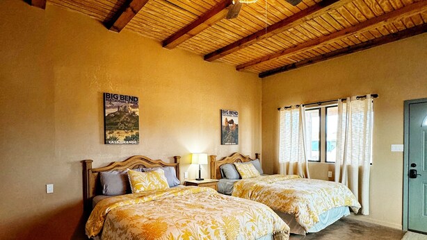 A bedroom with two beds, wooden ceiling beams, a ceiling fan, and posters on the wall. - Big Bend Bungalow A (TERLINGUA)