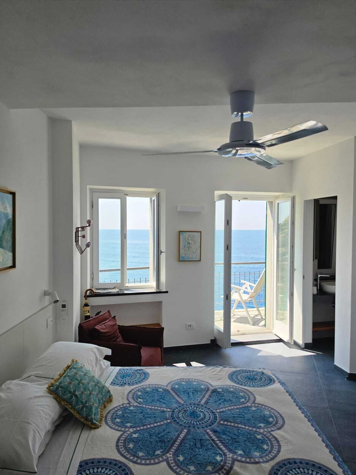 A bedroom with a bed, a ceiling fan, a window with a view of the sea, and an open door leading to a balcony.