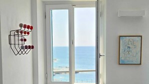 Een kamer met een raam dat uitkijkt over de zee, een rode bank, een wandrek met rode en grijze objecten en een ingelijste afbeelding.