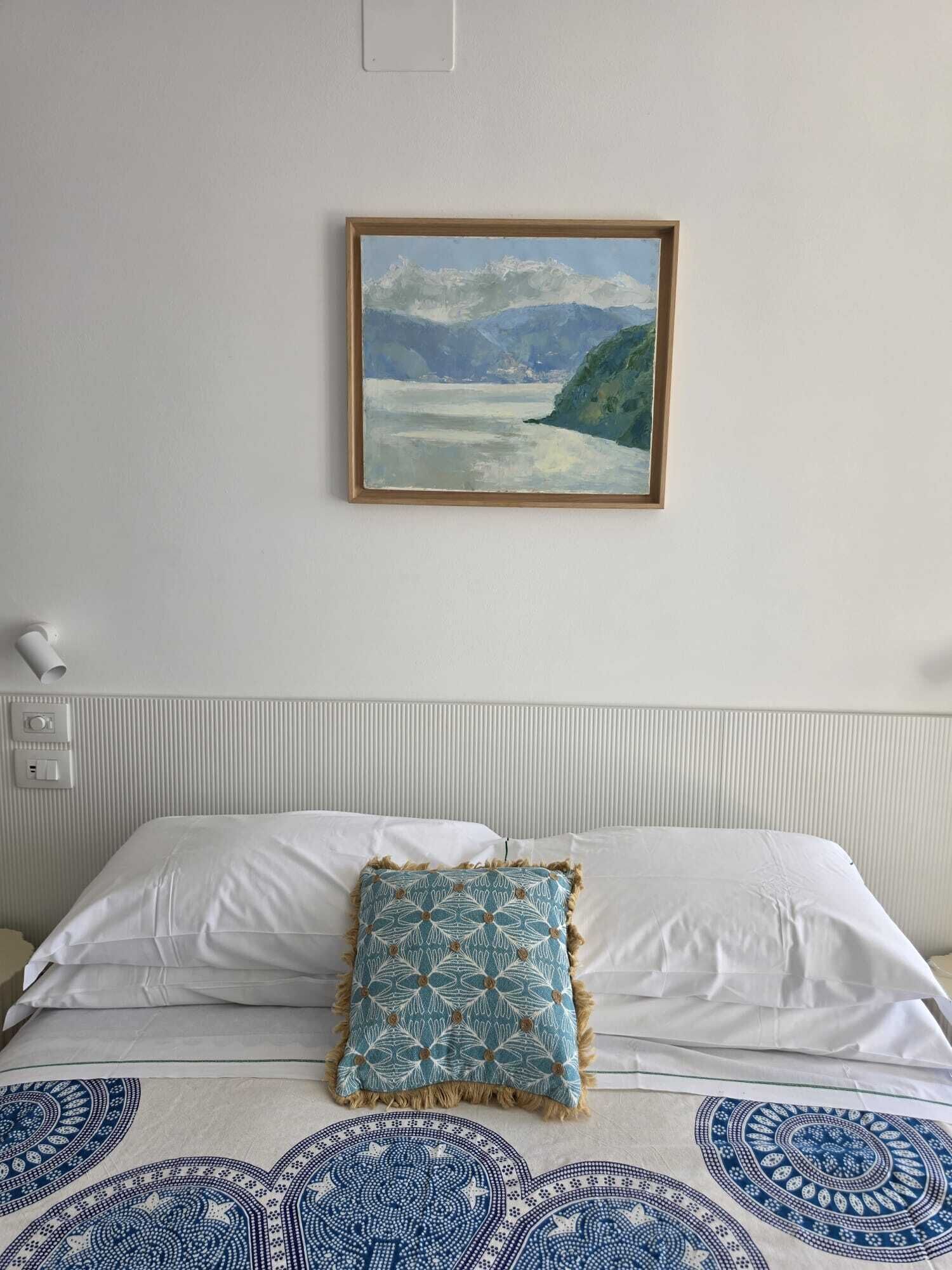 Een bed met witte lakens en een kussen met een patroon, een schilderij van een kustlandschap, en een witte muur met een lichtschakelaar en een lamp.
