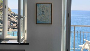 Een kamer met uitzicht op zee door open ramen, een ingelijste afbeelding aan de muur en een witte stoel buiten.