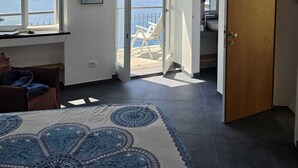 Een kamer met een plafondventilator, uitzicht op zee door open deuren, een houten deur en een kamerkleed met een patroon.