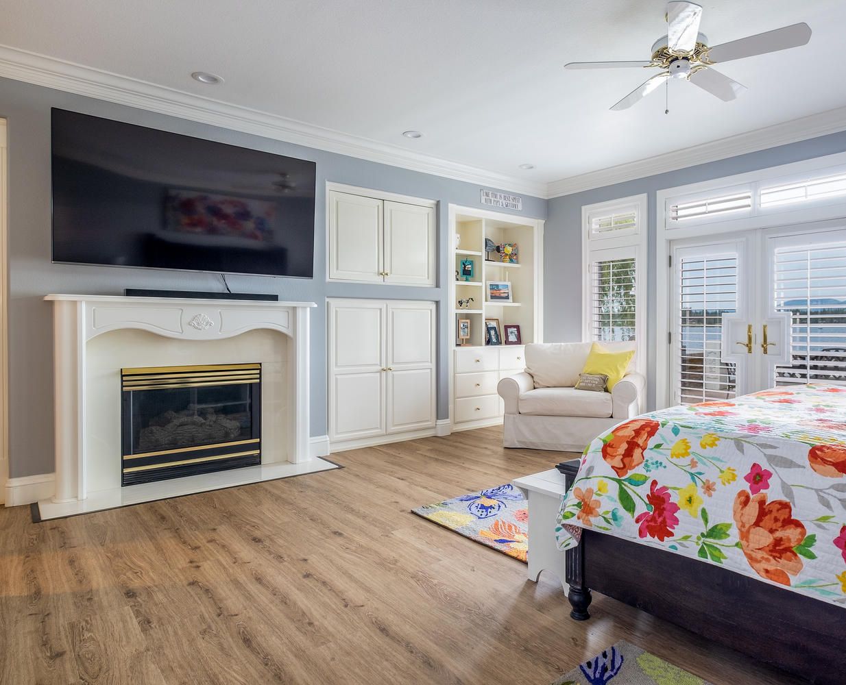 A bedroom with a fireplace, a bed with a floral bedspread, a television, and a view of the outdoors through large windows.
