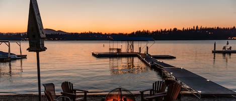 Un entorno junto al lago con una hoguera y sillas, un muelle que se adentra en el agua, y una vista despejada del atardecer sobre el agua.