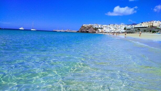 A beach with clear turquoise water, a white building with balconies, and a rocky outcrop. - Apartments Palm Garden (Morro Jable)