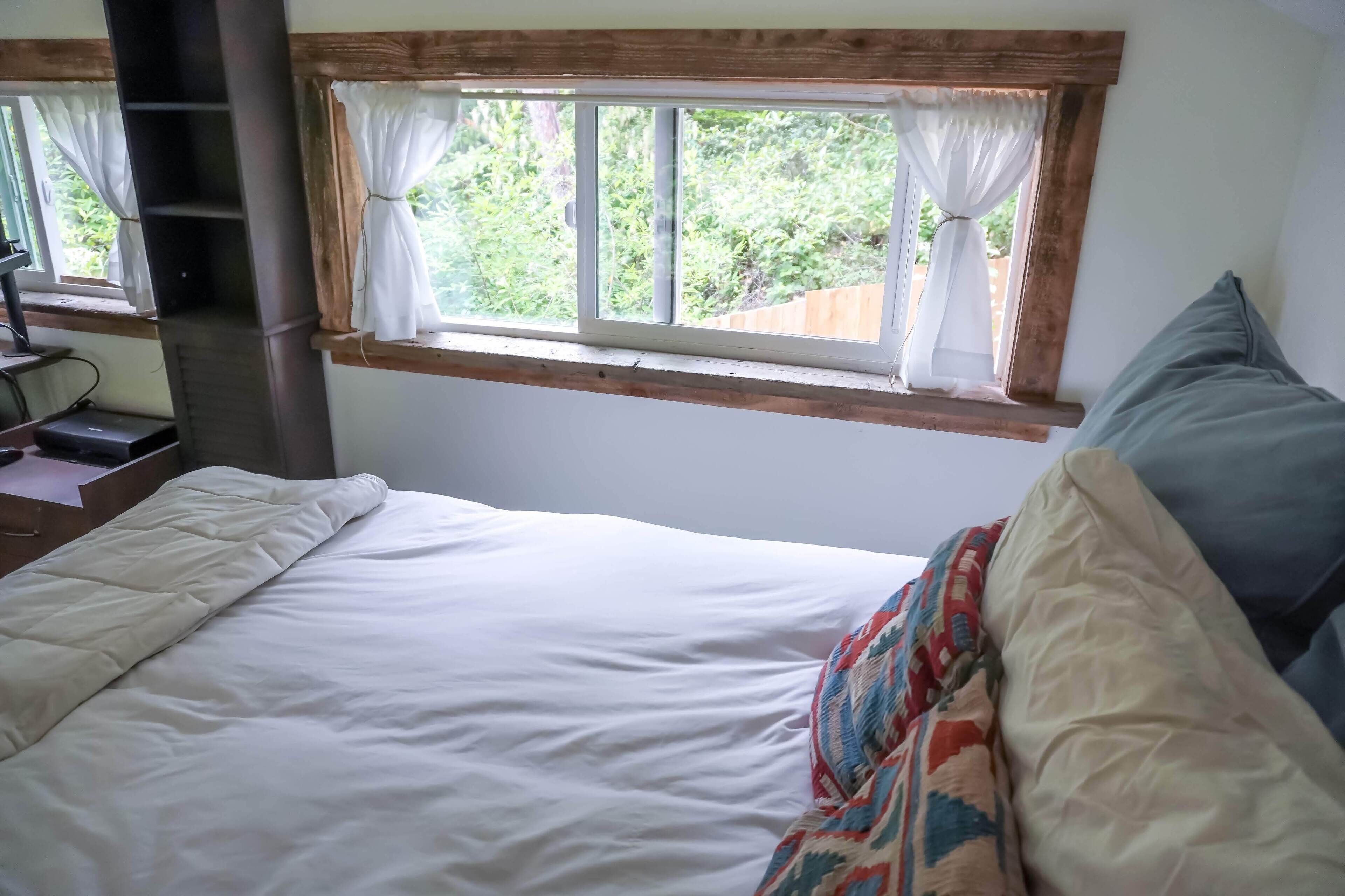 A bed with white bedding and a colorful blanket, a window with curtains, and a wooden shelf.