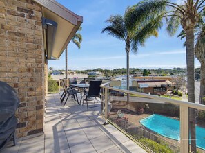 A balcony with a table and chairs, a pool, and palm trees. - Batehaven Coastal Escape | Pool & Bay Views (BATEHAVEN)