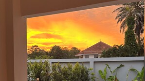 A room with a view of a sunset over a pool and garden, two chairs, and a bottle and glass on the windowsill. - Aonang Nine Poolvilla (Krabi)