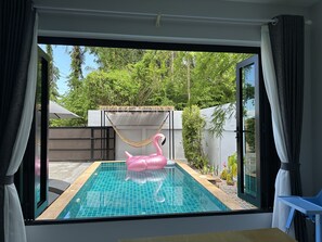 Blick auf einen Swimmingpool mit einem Flamingo-Schwimmspielzeug, umgeben von einer Holzterrasse und einer Struktur mit einem Reetdach.