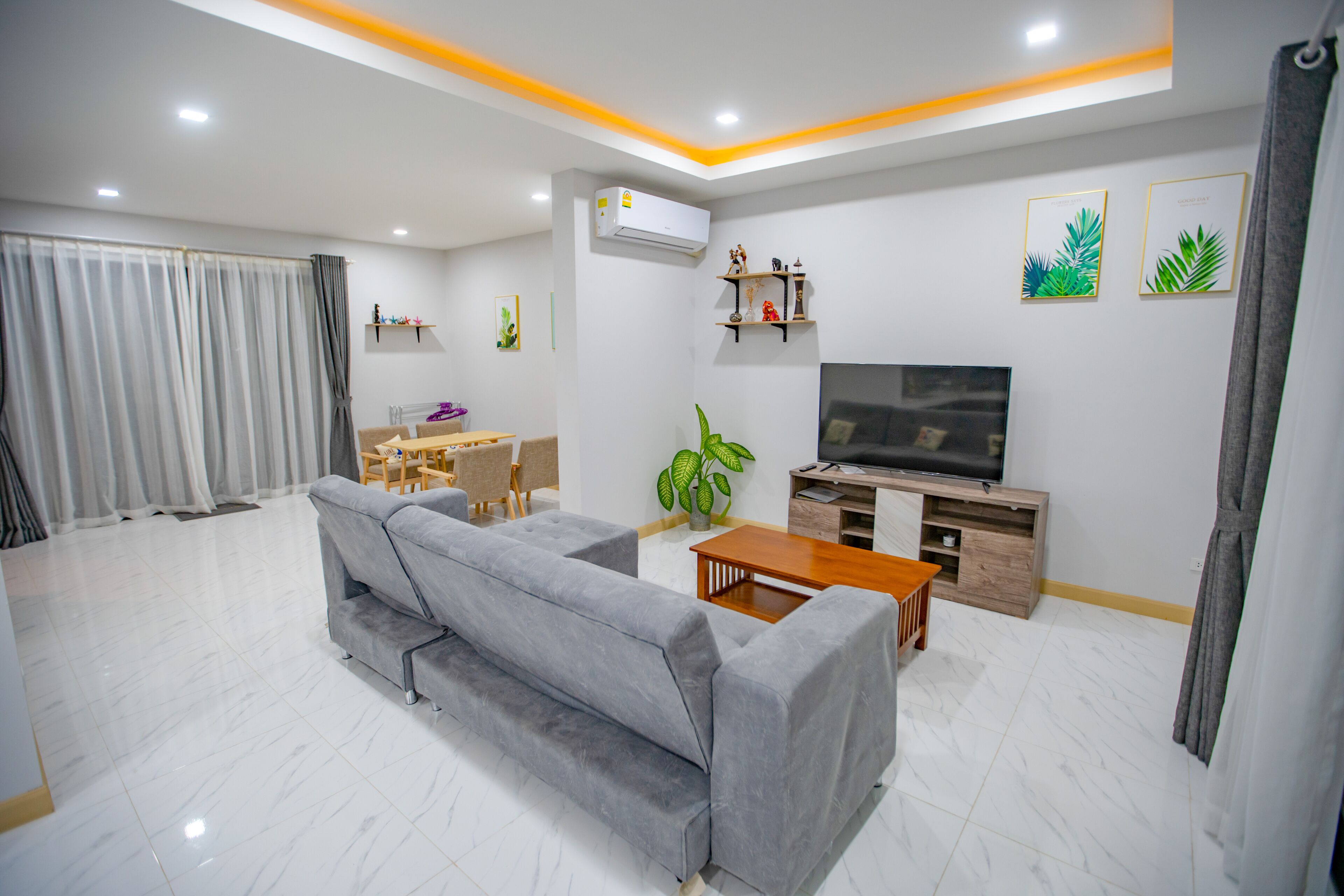 A modern living room with a grey sofa, wooden coffee table, and a TV on a wooden stand. There is a dining area with a table and chairs, and a potted plant.