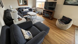 A living room with a sofa, coffee table, TV stand, and a wicker chair. - Riverwalk Condominiums #11 – Ground Floor with Private Patio, Close to Beach and (Seaside)