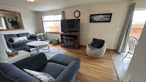 A living room with a sofa, a coffee table, a TV, and a wicker chair. - Riverwalk Condominiums #11 – Ground Floor with Private Patio, Close to Beach and (Seaside)