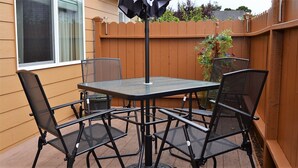 A patio with a square table and four chairs, surrounded by a wooden fence and a window. - Riverwalk Condominiums #11 – Ground Floor with Private Patio, Close to Beach and (Seaside)