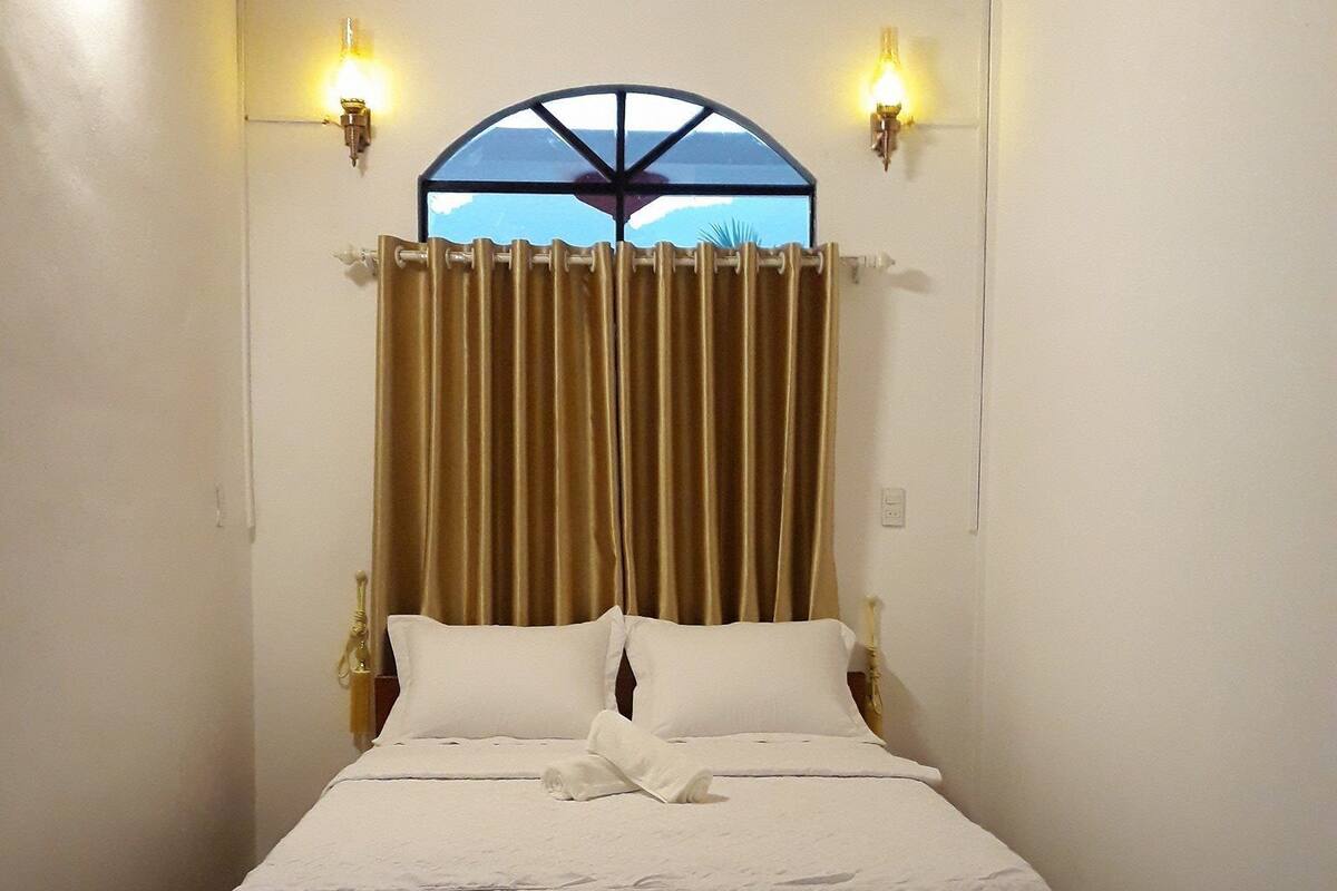 A neatly made bed with white linens, a red rug, and two wall-mounted lamps.