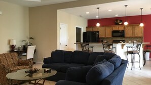A living room with a dark blue sectional sofa, a glass coffee table, and patterned armchairs. A kitchen with wooden cabinets and a red accent wall is visible in the background. - The Seahorse- 7 Hac (Laguna Vista)