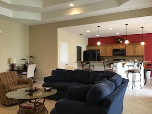 A living room with a dark blue sectional sofa, a glass coffee table, and patterned armchairs. A kitchen with wooden cabinets and a red accent wall is visible in the background.