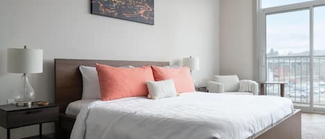 A modern bedroom with a large bed, two bedside tables with lamps, a cityscape painting, and a large window with a view.